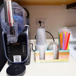 Puede incluir: Una máquina de granizados Ninja azul y negra con un código QR en el lateral. Una estación de granizados de colores arcoíris con vasos de plástico transparentes y pajitas de colores.