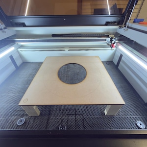 Puede incluir: Una máquina de corte por láser con una pieza de madera marrón claro en el interior. La pieza de madera tiene un gran recorte circular en el centro y está soportada por cuatro patas pequeñas. La máquina tiene una parte superior transparente y luces interiores brillantes.