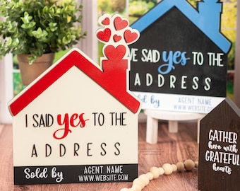 May include: Two wooden house-shaped signs with the text "I said yes to the address" and "We said yes to the address". Both signs have red roofs and hearts coming out of the chimney. A smaller wooden sign with the text "Gather here with grateful hearts" is in the background.