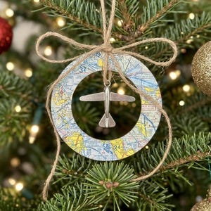 May include: A circular Christmas ornament featuring a colorful aviation map print with a silver-toned airplane charm suspended in the center. The ornament is tied with a natural brown jute twine bow at the top for hanging on a tree branch.