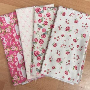 May include: Four fabric swatches with floral patterns. The fabrics feature pink roses, birds, and geometric designs on white and pink backgrounds. The fabrics are arranged on a light brown wooden surface.