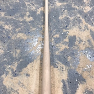 May include: A plain wooden baseball bat with a rounded end. The bat is lying on a light brown surface with gray patches.