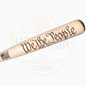 May include: A wooden baseball bat with the words "We the People" engraved in a decorative font. The bat is light-colored with a darker brown end. The words "Made in U.S.A." and a small American flag are printed near the handle.
