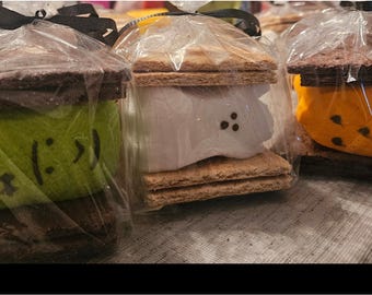 May include: Three individually wrapped s'mores treats featuring chocolate graham crackers sandwiching colorful marshmallow shapes. One green Frankenstein-style, one white ghost, and one orange pumpkin, each with hand-drawn black facial features.