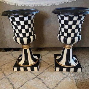 May include: Two decorative urns with a black and white checkered pattern on the body. The urns have a black rim, a gold accent, and a square base. The base and the body have a black and white striped pattern.