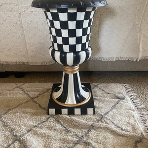 May include: Black and white checkered ceramic planter with a gold trim. The planter has a wide base and a narrow top. It is sitting on a beige rug.