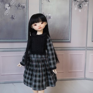 May include: A doll wearing a black and white plaid dress with a black top. The dress has long sleeves and a pleated skirt. The doll has long black hair and is standing in front of a light pink wall.