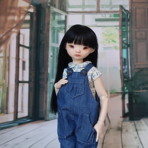 May include: A ball-jointed doll with long, straight black hair and a fringe, wearing a white short-sleeved shirt with a small blue floral pattern underneath dark blue denim dungarees that feature a front bib pocket and two side pockets.