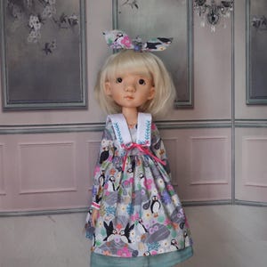 May include: A doll with blonde hair and a patterned dress featuring puffins, flowers, and sea elements. The dress has a white collar with blue embroidery and a pink ribbon. The doll wears a matching bow in her hair.
