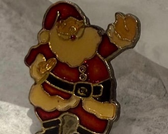 May include: A festive Santa Claus pin with a red and gold color scheme. The pin features a jolly Santa Claus design with a red hat and suit, a gold beard, and black boots. The pin is made of metal and has a shiny finish.