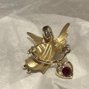 Könnte beinhalten: Eine goldfarbene Engelsbrosche mit ausgebreiteten Flügeln und einem herzförmigen Anhänger. Der Herzanhänger hat einen roten Edelstein. Der Engel ist in einem fließenden Gewand dargestellt.