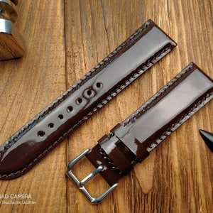 Pu&ograve; includere: Due cinturini per orologio in pelle marrone con cuciture bianche. I cinturini sono lucidi e hanno una finitura lucida. I cinturini sono disposti su una superficie di legno.