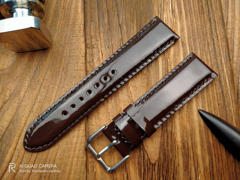 Pu&ograve; includere: Due cinturini per orologio in pelle marrone con cuciture bianche. I cinturini sono lucidi e hanno una finitura lucida. I cinturini sono disposti su una superficie di legno.