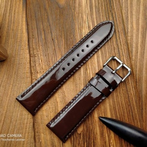 Pu&ograve; includere: Un cinturino da orologio in pelle marrone con finitura lucida. Il cinturino ha una fibbia argentata ed &egrave; mostrato su una superficie di legno.