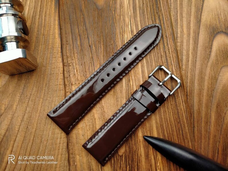 Pu&ograve; includere: Un cinturino da orologio in pelle marrone con finitura lucida. Il cinturino ha una fibbia argentata ed &egrave; mostrato su una superficie di legno.