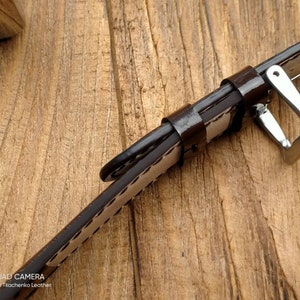 Pu&ograve; includere: Un cinturino da orologio in pelle marrone con cuciture bianche e una fibbia argentata. Il cinturino &egrave; appoggiato su una superficie di legno.