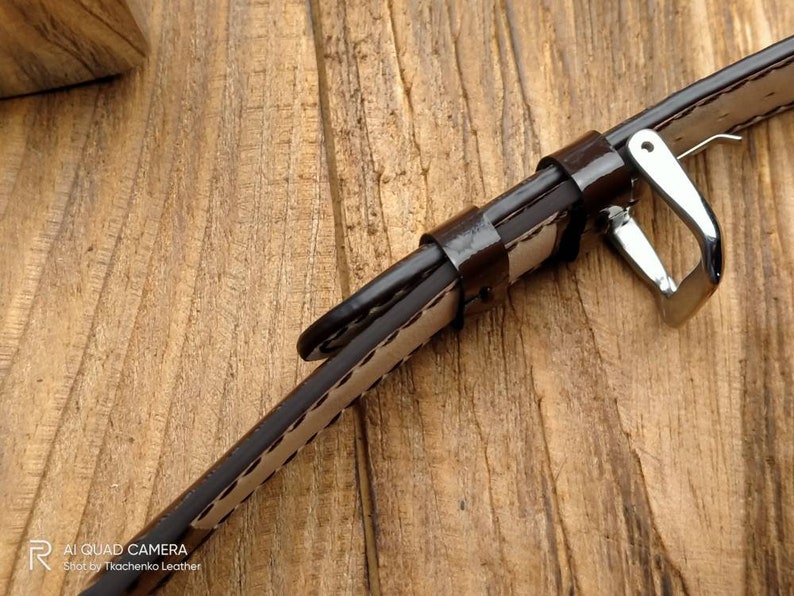 Pu&ograve; includere: Un cinturino da orologio in pelle marrone con cuciture bianche e una fibbia argentata. Il cinturino &egrave; appoggiato su una superficie di legno.