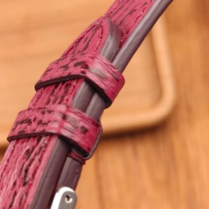 May include: Close-up of a burgundy watch strap with a textured, patterned surface. The strap features a dark brown border and stitching, with a silver-colored buckle. The watch strap is made of a durable material.