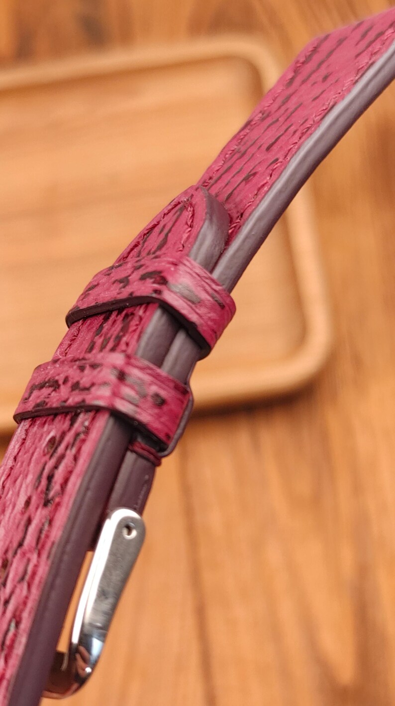 May include: Close-up of a burgundy watch strap with a textured, patterned surface. The strap features a dark brown border and stitching, with a silver-colored buckle. The watch strap is made of a durable material.