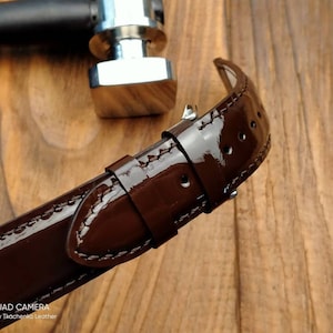 Pu&ograve; includere: Un cinturino da orologio in pelle marrone con finitura lucida. Il cinturino ha cuciture bianche ed &egrave; mostrato su una superficie di legno.