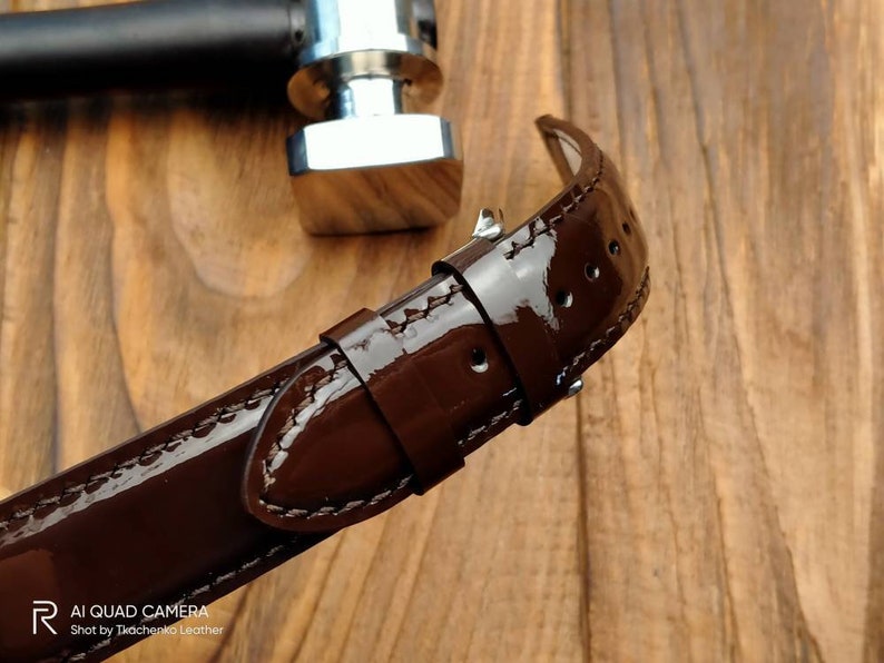 Pu&ograve; includere: Un cinturino da orologio in pelle marrone con finitura lucida. Il cinturino ha cuciture bianche ed &egrave; mostrato su una superficie di legno.