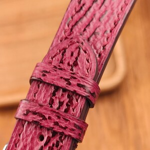 May include: Close-up of a vibrant red watch strap with a textured, patterned surface. The strap features a buckle and multiple adjustment holes. The leather band has a unique, eye-catching design.