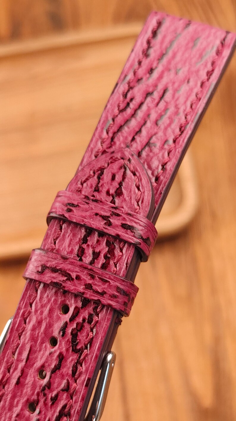 May include: Close-up of a vibrant red watch strap with a textured, patterned surface. The strap features a buckle and multiple adjustment holes. The leather band has a unique, eye-catching design.
