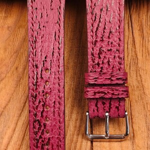 May include: Two burgundy watch straps with a textured surface and a silver-colored buckle. The straps are made of a material that resembles sharkskin. The background is a wooden surface.