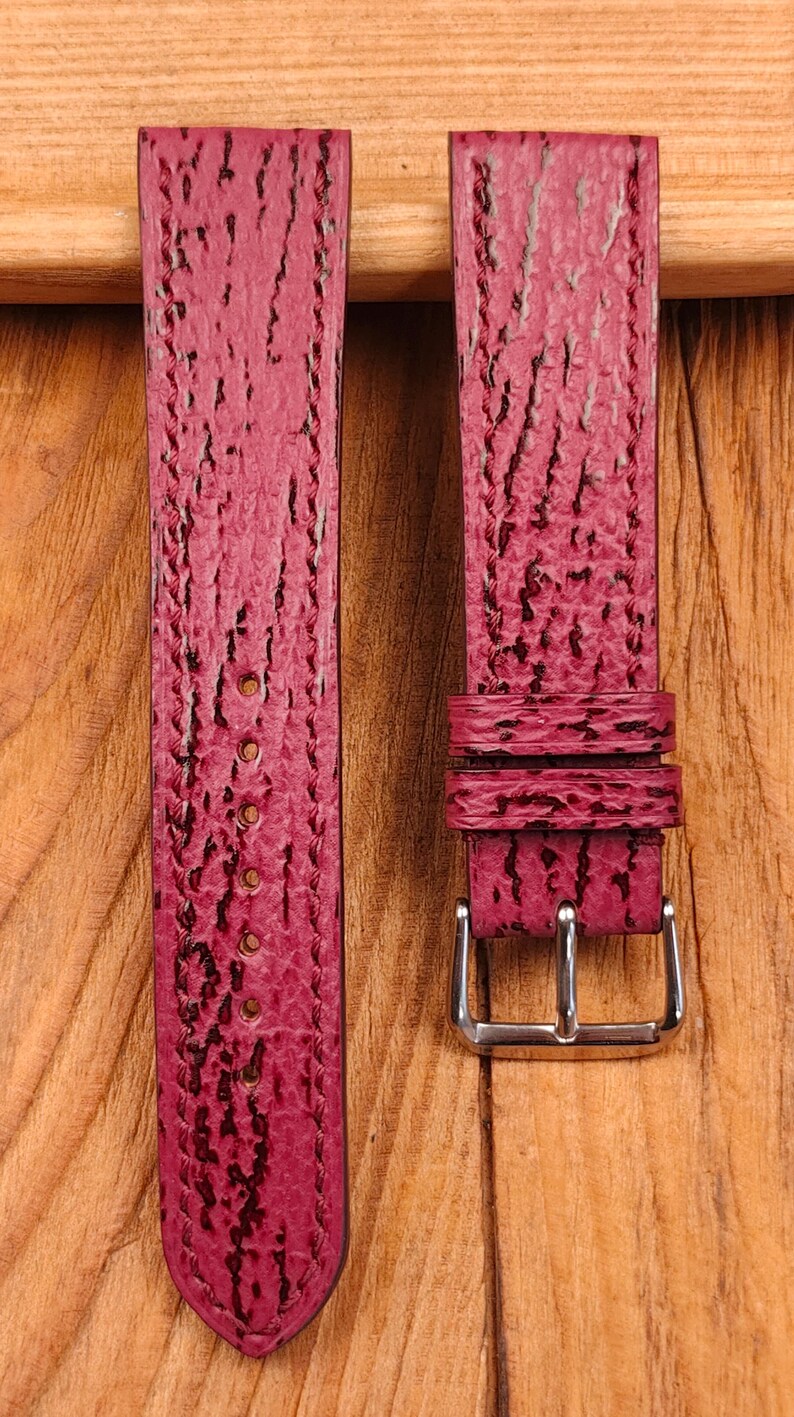 May include: Two burgundy watch straps with a textured surface and a silver-colored buckle. The straps are made of a material that resembles sharkskin. The background is a wooden surface.