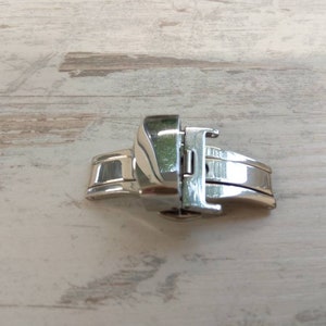 May include: A close-up of a shiny, silver watch clasp. The clasp is made of polished metal and has a secure, folding design. The watch clasp is designed to fasten a watch band.