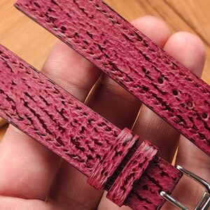 May include: Close-up of a burgundy watch strap with a textured pattern and a silver buckle. The strap is made of a material with a dark, linear design. The watch band is shown against a wooden background.