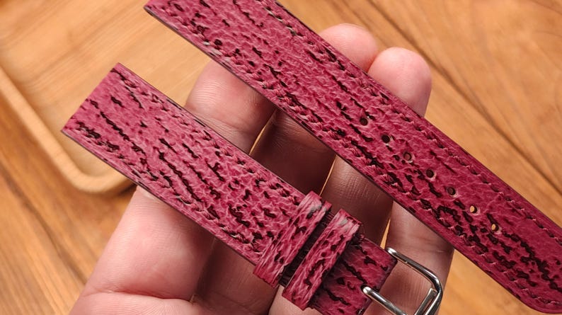 May include: Close-up of a burgundy watch strap with a textured pattern and a silver buckle. The strap is made of a material with a dark, linear design. The watch band is shown against a wooden background.