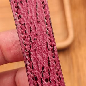 May include: Close-up of a watch strap in a deep pink hue with a textured surface and black accents. The edges are stitched with pink thread. A silver buckle is visible.