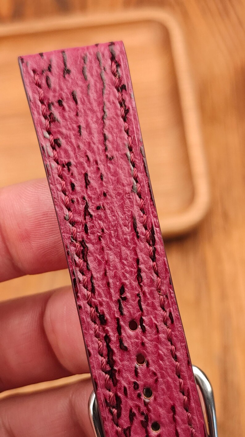 May include: Close-up of a watch strap in a deep pink hue with a textured surface and black accents. The edges are stitched with pink thread. A silver buckle is visible.