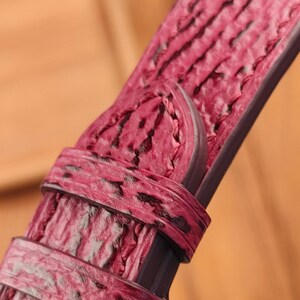 May include: Close-up of a watch strap in a deep pink hue with a textured, patterned surface. The strap features a buckle and a loop, showcasing the intricate details of the material and design. The strap is a rich, saturated color.