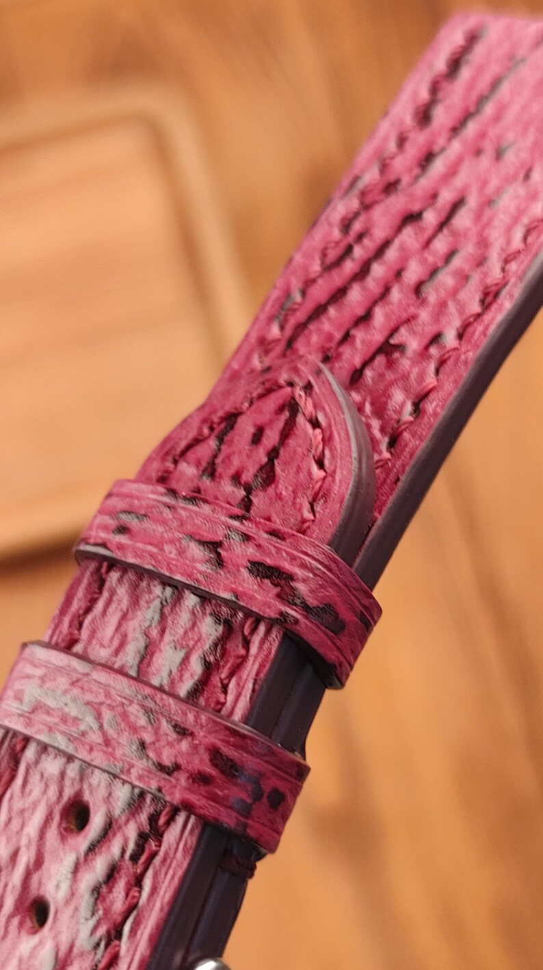 May include: Close-up of a watch strap in a deep pink hue with a textured, patterned surface. The strap features a buckle and a loop, showcasing the intricate details of the material and design. The strap is a rich, saturated color.