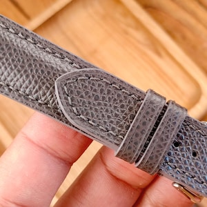 Puede incluir: Primer plano de una correa de reloj de cuero gris con una superficie texturizada y costuras en contraste. La correa tiene una hebilla y una presilla para asegurar el reloj. La correa es de color neutro.