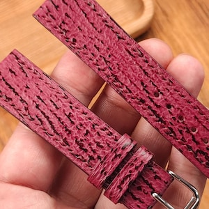 May include: Close-up of a burgundy watch strap with a textured pattern and a silver buckle. The strap is held in a hand, showcasing the detailed grain and color variations. The watch band is made of leather.