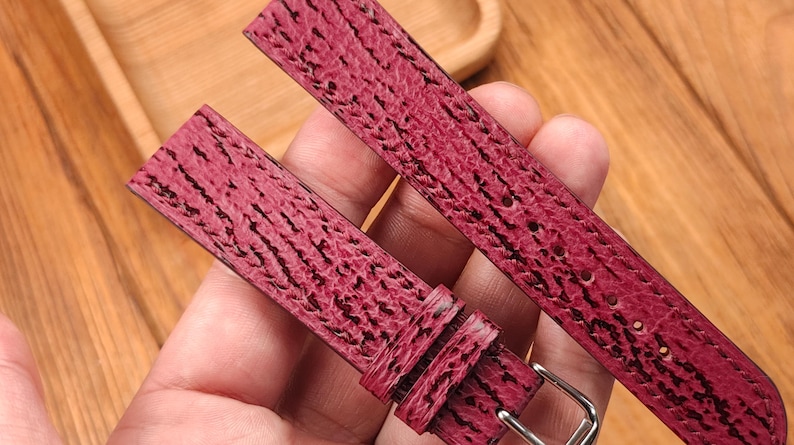 May include: Close-up of a burgundy watch strap with a textured pattern and a silver buckle. The strap is held in a hand, showcasing the detailed grain and color variations. The watch band is made of leather.