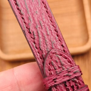 May include: Close-up of a burgundy leather watch strap with a textured surface and dark stitching. The strap has a buckle and a loop for securing the watch. The background is a wooden tray.