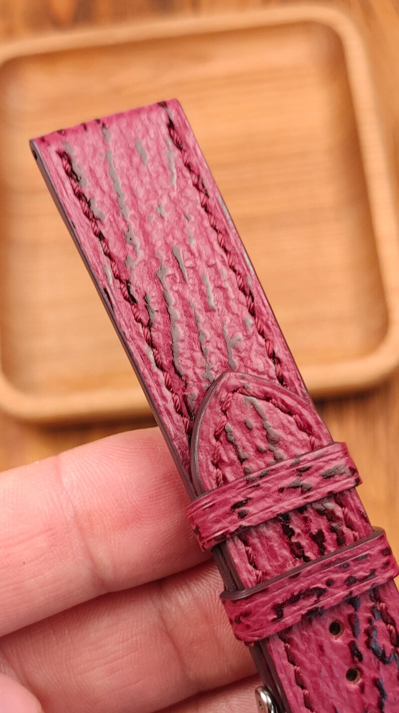 May include: Close-up of a burgundy leather watch strap with a textured surface and dark stitching. The strap has a buckle and a loop for securing the watch. The background is a wooden tray.