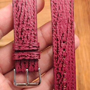 May include: A pair of burgundy watch straps with a textured surface and black accents. One strap has a silver buckle, and the other has several holes for adjustment. The straps are held against a wooden background.
