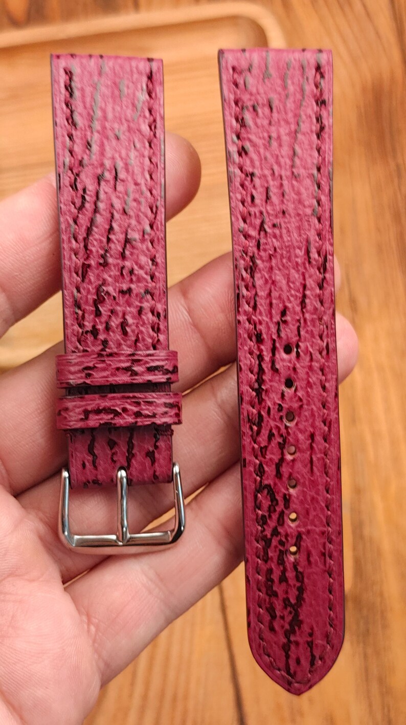 May include: A pair of burgundy watch straps with a textured surface and black accents. One strap has a silver buckle, and the other has several holes for adjustment. The straps are held against a wooden background.