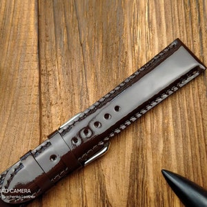 Pu&ograve; includere: Un cinturino da orologio in pelle marrone con finitura lucida e cuciture bianche. Il cinturino &egrave; su una superficie di legno.