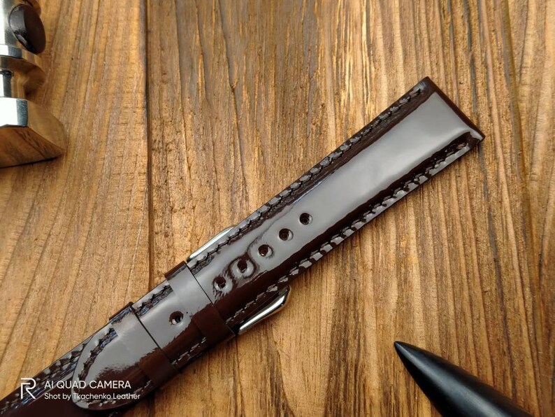 Pu&ograve; includere: Un cinturino da orologio in pelle marrone con finitura lucida e cuciture bianche. Il cinturino &egrave; su una superficie di legno.