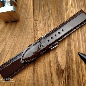 Pu&ograve; includere: Un cinturino da orologio in pelle marrone scuro con finitura lucida e cuciture bianche. Il cinturino &egrave; appoggiato su una superficie in legno.