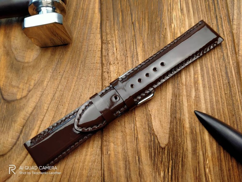 Pu&ograve; includere: Un cinturino da orologio in pelle marrone scuro con finitura lucida e cuciture bianche. Il cinturino &egrave; appoggiato su una superficie in legno.