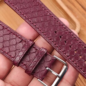 May include: Burgundy watch strap with a textured, scale-like pattern. The strap features a silver-colored buckle and is made of a material that resembles snakeskin. The stitching is a matching burgundy color.