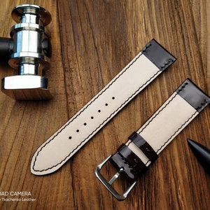 Pu&ograve; includere: Due cinturini per orologio in pelle marrone con cuciture bianche. I cinturini hanno una finitura lucida e sono mostrati su una superficie in legno.