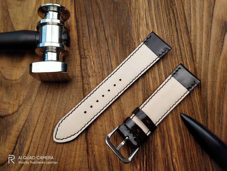 Pu&ograve; includere: Due cinturini per orologio in pelle marrone con cuciture bianche. I cinturini hanno una finitura lucida e sono mostrati su una superficie in legno.